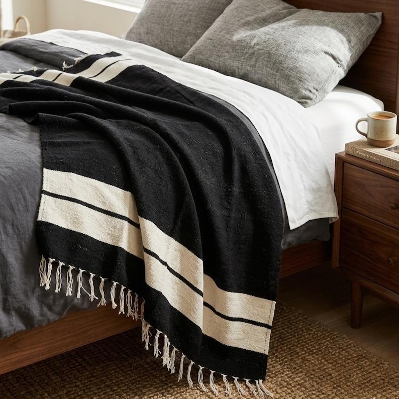 Puede incluir: Una manta a rayas blancas y negras con flecos, sobre una cama. La manta presenta dos anchas rayas blancas sobre un fondo negro. La cama tiene ropa de cama y almohadas grises, con una mesita de noche de madera y una taza.