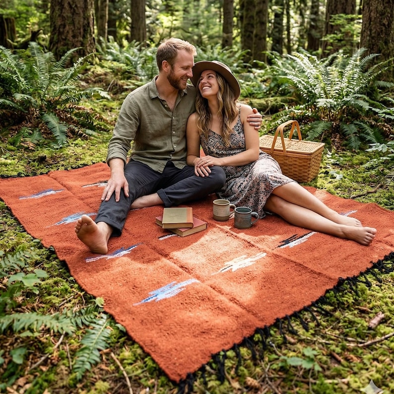 Pu&ograve; includere: Una coperta da picnic tessuta color ruggine con frange nere, con un design geometrico con accenti blu e bianchi. La coperta &egrave; stesa su un pavimento forestale muschioso, con un cestino da picnic e tazze nelle vicinanze.