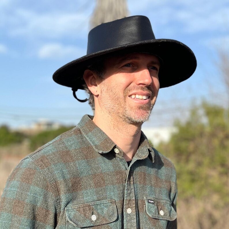 May include: A black hat with a wide brim and a tall crown. The hat is worn by a person wearing a plaid shirt with shades of blue and brown. The background shows a blurred outdoor scene with a blue sky.