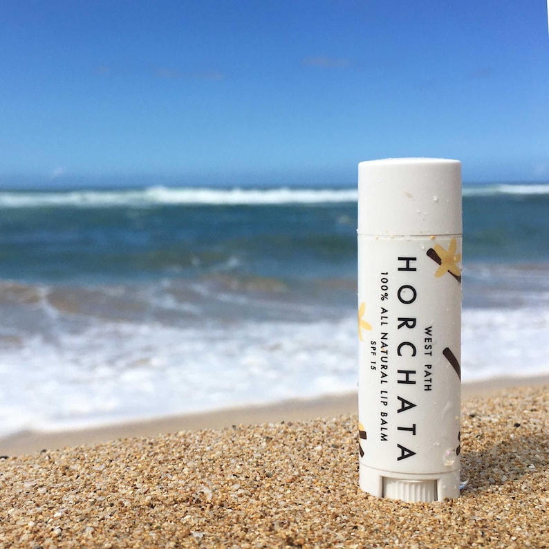 Puede incluir: Un tubo de b&aacute;lsamo labial blanco con el texto "HORCHATA" y "100% ALL NATURAL LIP BALM SPF 15" est&aacute; en una playa de arena con el oc&eacute;ano al fondo. El b&aacute;lsamo labial tiene un gr&aacute;fico de vainilla y la marca "WEST PATH".