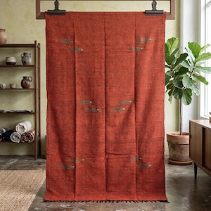 May include: A large, rust-coloured textile with a textured appearance, hanging from a wooden frame. The textile features embroidered accents in a muted green colour. The bottom edge has a black fringe. The textile is displayed in a well-lit room with a neutral background.