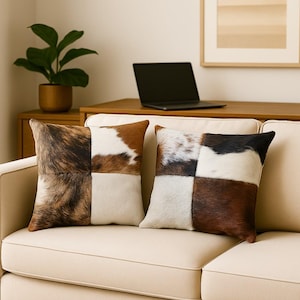 May include: Two square decorative throw pillows with a patchwork design. Each pillow features four sections of cowhide in shades of brown, white, and black. The pillows are placed on a cream-colored sofa.