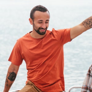 K&ouml;nnte beinhalten: Ein rostfarbenes T-Shirt mit kurzen &Auml;rmeln. Das Shirt besteht aus einem weichen Material und hat einen Rundhalsausschnitt. Die Person, die das Shirt tr&auml;gt, l&auml;chelt. Der Hintergrund ist ein Gew&auml;sser.