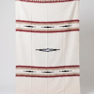 May include: A rectangular woven blanket in off-white featuring horizontal stripes in deep red and tan. The design includes a series of black geometric diamond-shaped motifs across the center and near the top and bottom edges, finished with fringe.