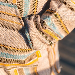 May include: Close-up of a beige fabric with a herringbone pattern. The fabric features stripes in yellow, teal, and black. The sleeve of a garment is visible, with a folded cuff. A small yellow tag is attached to the garment.