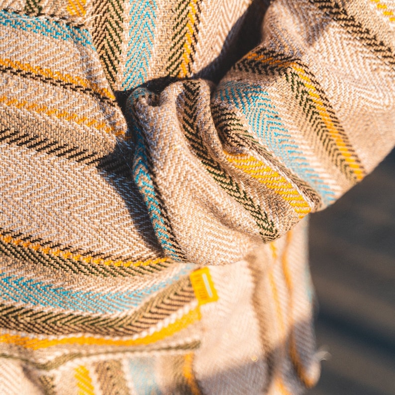 May include: Close-up of a beige fabric with a herringbone pattern. The fabric features stripes in yellow, teal, and black. The sleeve of a garment is visible, with a folded cuff. A small yellow tag is attached to the garment.