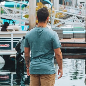May include: A person wearing a teal t-shirt and khaki shorts stands near water. The person has tattoos on their arms and neck. A boat and dock are visible in the background. The t-shirt has short sleeves.