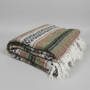 May include: A folded woven blanket featuring a striped pattern in shades of sage green, earthy brown, and cream. The textile displays a textured weave with black geometric accents and long, white fringe detailing along the finished edge.