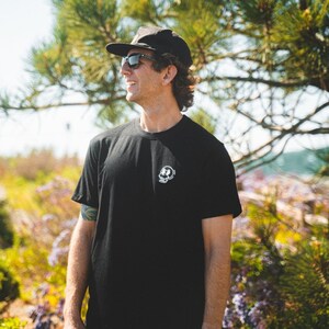 May include: Black crew neck t-shirt with a small white graphic on the chest. The graphic appears to be a stylized skull design. The person in the image is wearing a black cap and sunglasses.