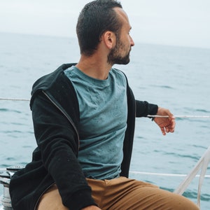 May include: A person wearing a blue t-shirt and a black zip-up hoodie, with tan trousers, sitting on a boat. The background is the sea and a cloudy sky. The person is looking out at the water.