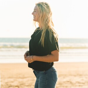 K&ouml;nnte beinhalten: Ein schwarzes T-Shirt mit kurzen &Auml;rmeln und eine blaue Jeans. Die Person steht an einem Sandstrand mit dem Meer im Hintergrund. Die Person l&auml;chelt und blickt auf das Meer.
