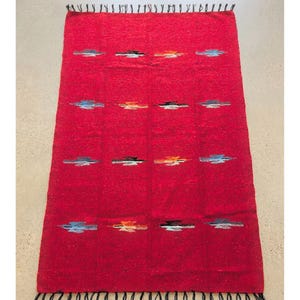 May include: A rectangular red rug with a woven texture. The rug features rows of colorful horizontal designs in blue, black, and orange. The edges are finished with black fringe.