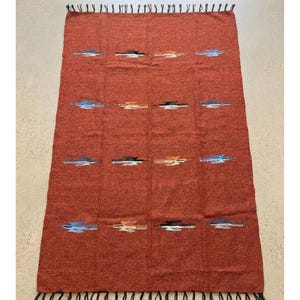Pu&ograve; includere: Un tappeto rettangolare tessuto color ruggine con una superficie strutturata. Il tappeto presenta un motivo ripetuto di linee orizzontali blu, bianche, nere e arancioni. I bordi sono rifiniti con frange.