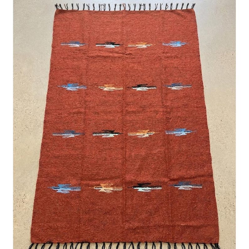 Pu&ograve; includere: Un tappeto rettangolare tessuto color ruggine con una superficie strutturata. Il tappeto presenta un motivo ripetuto di linee orizzontali blu, bianche, nere e arancioni. I bordi sono rifiniti con frange.
