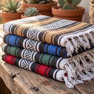 May include: A stack of four woven blankets featuring a repeating geometric pattern in black and white, with solid color bands in mustard yellow, royal blue, forest green, and bright red. Each blanket has white fringe trim along the short edges.