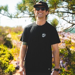 May include: Black crew neck t-shirt with a small white graphic on the chest. The graphic appears to be a skull with a circular design around it. The person in the image is wearing a black cap and sunglasses.