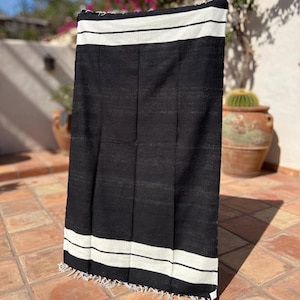 Puede incluir: Una manta tejida en blanco y negro con un dise&ntilde;o a rayas. La manta tiene un tejido grueso y texturizado y una franja a lo largo del borde inferior. Las rayas blancas est&aacute;n en la parte superior e inferior de la manta.