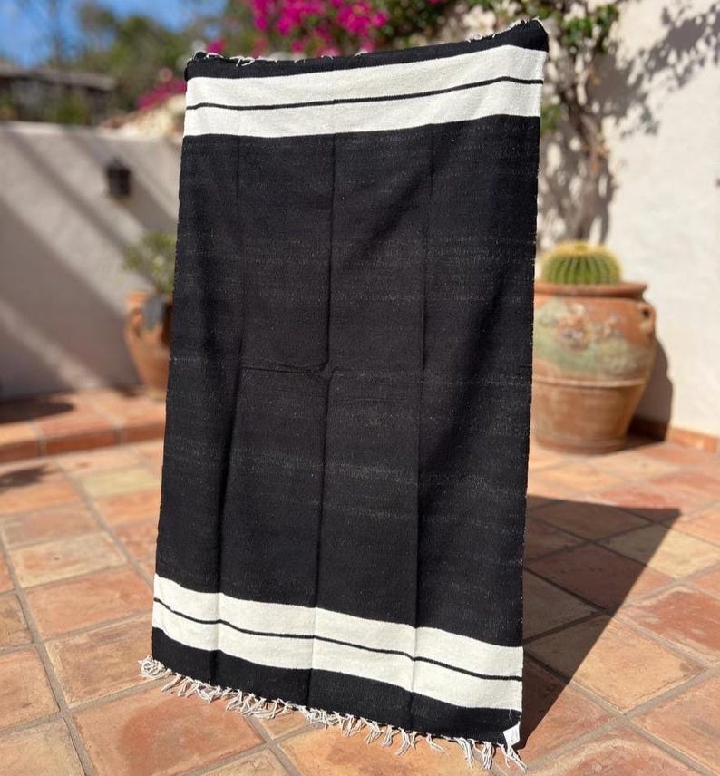 Puede incluir: Una manta tejida en blanco y negro con un dise&ntilde;o a rayas. La manta tiene un tejido grueso y texturizado y una franja a lo largo del borde inferior. Las rayas blancas est&aacute;n en la parte superior e inferior de la manta.