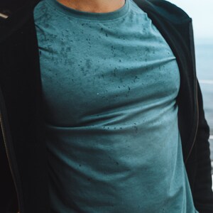 May include: A teal crew neck t-shirt with a speckled design. The shirt is worn under a black jacket. The image is a medium shot, focusing on the upper body and clothing. The t-shirt has a worn look.