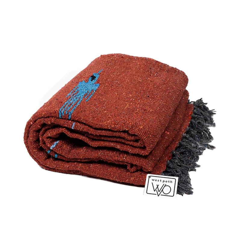 Pu&ograve; includere: Coperta piegata color ruggine con un disegno blu e frange grigie. La coperta &egrave; realizzata in un materiale strutturato e presenta un'etichetta rettangolare bianca con le parole "west path" e le lettere "WO" in un font stilizzato.