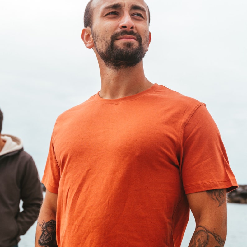 K&ouml;nnte beinhalten: Ein orangefarbenes T-Shirt mit Rundhalsausschnitt. Das kurz&auml;rmelige Shirt ist aus einem weichen Material. Die Person, die das Shirt tr&auml;gt, hat einen Bart und T&auml;towierungen am Arm. Der Hintergrund ist ein bew&ouml;lkter Himmel und Wasser.