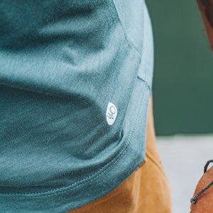 May include: Close-up of a teal t-shirt with a small white logo. The shirt features a curved hem and is made of a soft, textured fabric. The t-shirt is paired with tan trousers. The image focuses on the shirt's texture and design.