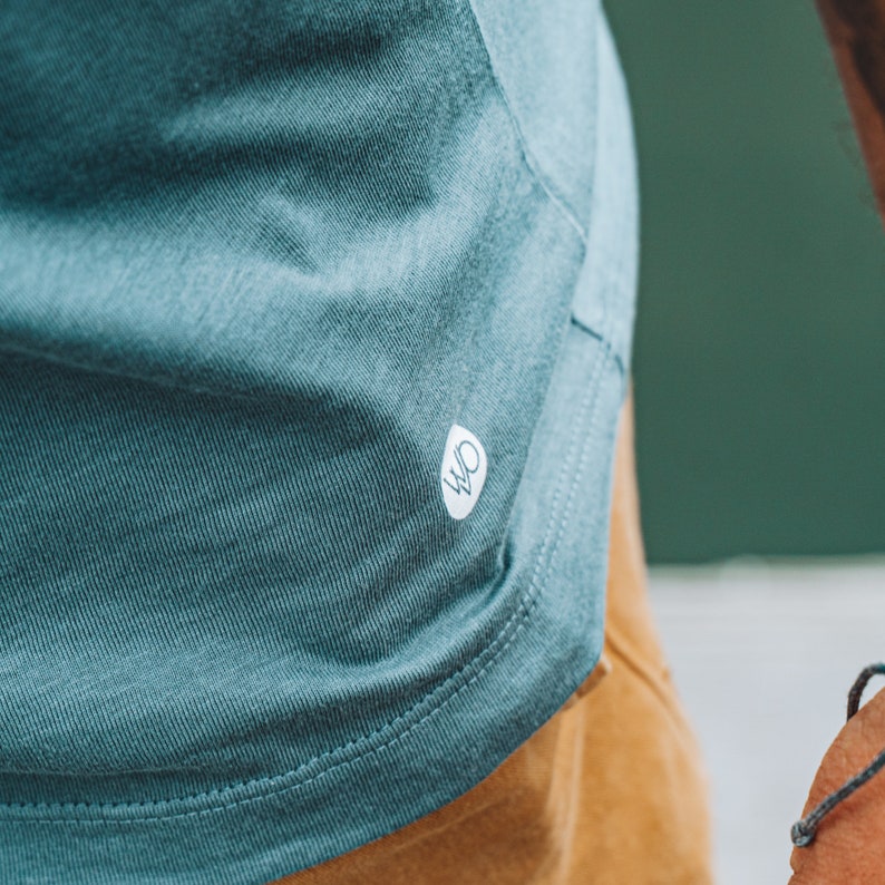 May include: Close-up of a teal t-shirt with a small white logo. The shirt features a curved hem and is made of a soft, textured fabric. The t-shirt is paired with tan trousers. The image focuses on the shirt's texture and design.