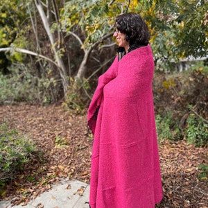 May include: A person wearing a pink fuzzy blanket with black trim. The person is standing in a wooded area with fallen leaves on the ground.