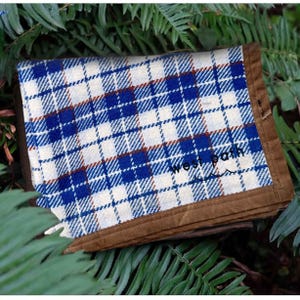 May include: A folded, square fabric item with a blue and white plaid pattern and brown trim. The text "west path" is printed on the fabric. The item is resting on a bed of green leaves.