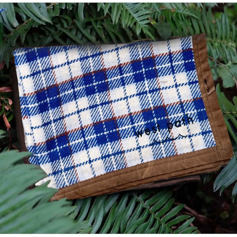 May include: A folded, square fabric item with a blue and white plaid pattern and brown trim. The text "west path" is printed on the fabric. The item is resting on a bed of green leaves.