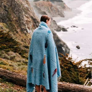 K&ouml;nnte beinhalten: Eine Person, eingeh&uuml;llt in eine blaue Decke mit bunten Mustern, steht auf einer Klippe mit Blick auf eine felsige K&uuml;ste. Die Person blickt auf das Meer.