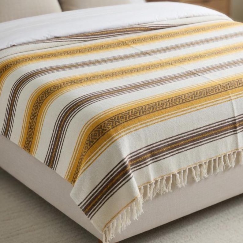 May include: A woven blanket with a striped pattern in cream, mustard yellow, and brown. The blanket has a fringe along the bottom edge. The design includes alternating stripes and decorative geometric patterns. This blanket is suitable for a bed or sofa.