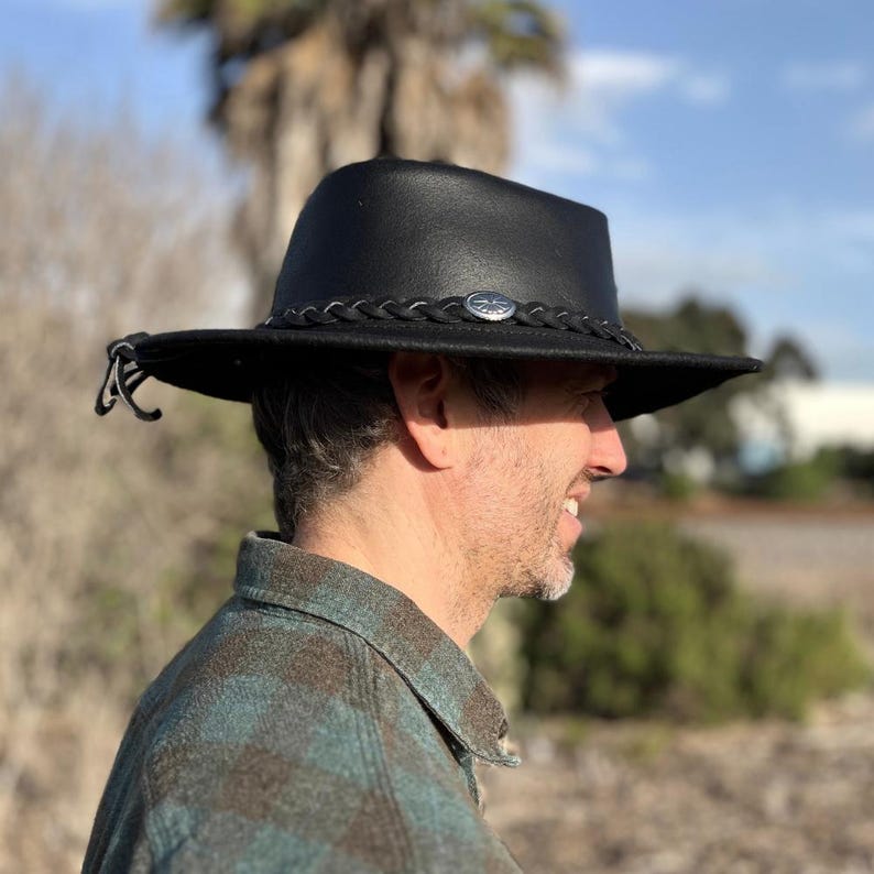 May include: Black leather hat with a braided band and a silver-colored emblem. The hat has a wide brim and a classic design. The hat is a stylish accessory for outdoor activities.