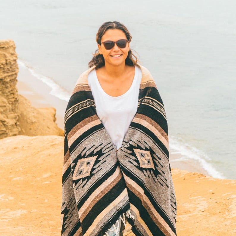 May include: A person wearing sunglasses and a white shirt is wrapped in a black, brown and grey striped blanket with a diamond pattern. The person is standing on a sandy beach with a body of water in the background.