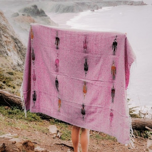 Könnte beinhalten: Eine rosa Decke mit einem sich wiederholenden Muster stilisierter Figuren in Schwarz, Orange und Rosa. Die Decke hat einen Fransenrand und wird vor einem Küstenhintergrund gehalten.