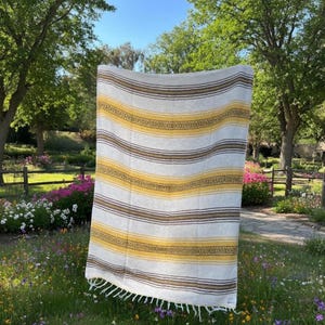 May include: A rectangular blanket with horizontal stripes in shades of white, yellow, and brown. The blanket has a fringe along the bottom edge. The blanket is hanging outdoors in a garden setting.