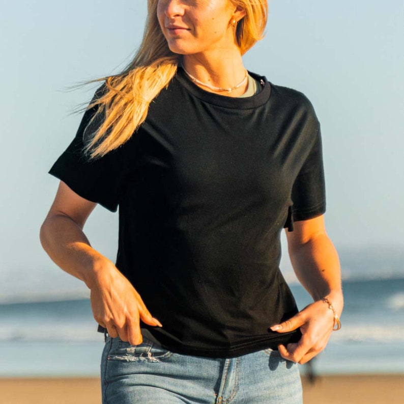 K&ouml;nnte beinhalten: Ein schwarzes T-Shirt mit Rundhalsausschnitt und kurzen &Auml;rmeln. Das Shirt wird mit einer blauen Jeans kombiniert. Das Model steht drau&szlig;en in der N&auml;he des Meeres an einem sonnigen Tag.