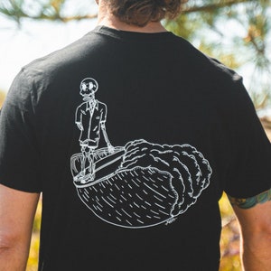 May include: Black t-shirt featuring a white graphic of a skeleton in a suit surfing a wave. The illustration is detailed and stylized, with the skeleton standing on a surfboard. The design is centered on the back of the shirt.