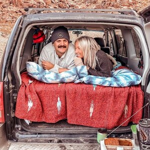 Pu&ograve; includere: Una coppia si rilassa nel retro di un veicolo, adagiata su una coperta rossa con un motivo bianco e blu. Sono circondati da cuscini e un sacco a pelo. Il portellone del veicolo &egrave; aperto, rivelando uno scenario esterno.