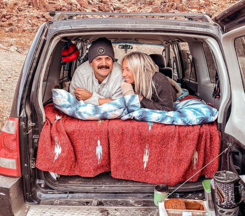 Pu&ograve; includere: Una coppia si rilassa nel retro di un veicolo, adagiata su una coperta rossa con un motivo bianco e blu. Sono circondati da cuscini e un sacco a pelo. Il portellone del veicolo &egrave; aperto, rivelando uno scenario esterno.