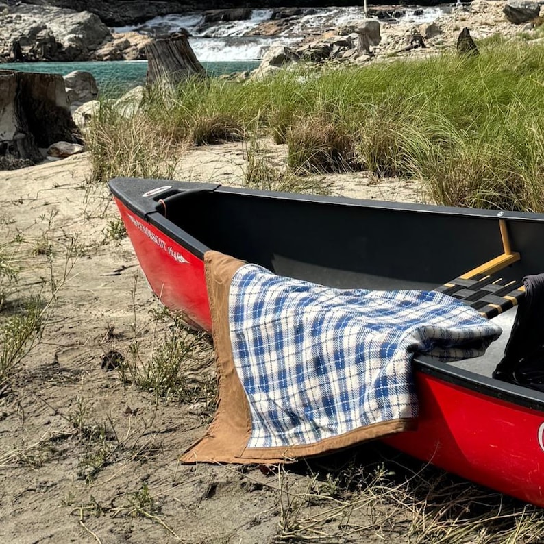 May include: A red canoe with a black interior rests on a sandy shore. A blue and white plaid blanket with a brown border is draped over the side. The background features a river and grassy area.