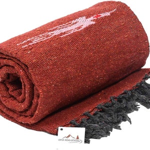Pu&ograve; includere: Coperta tessuta color terracotta arrotolata con un disegno bianco e rosa. La coperta ha una frangia nera lungo un bordo. Il logo Open Road Goods &egrave; visibile su una piccola etichetta.