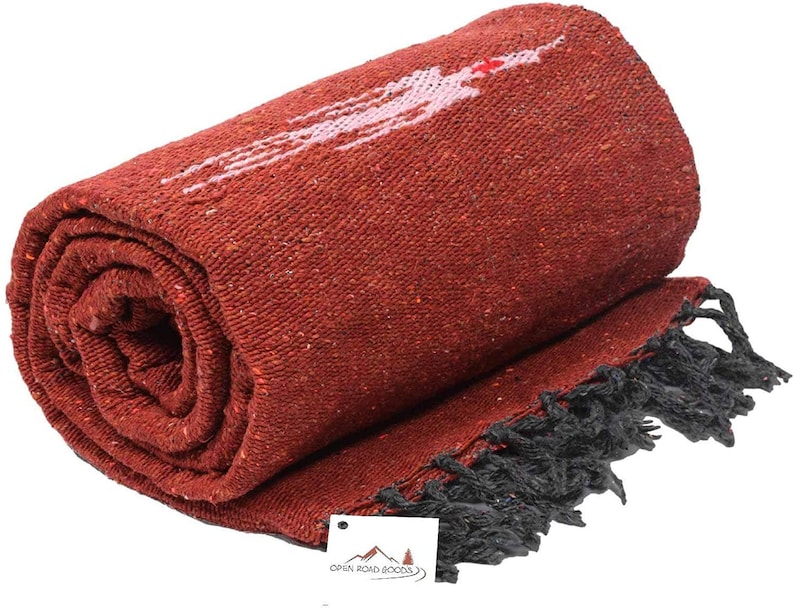 Pu&ograve; includere: Coperta tessuta color terracotta arrotolata con un disegno bianco e rosa. La coperta ha una frangia nera lungo un bordo. Il logo Open Road Goods &egrave; visibile su una piccola etichetta.