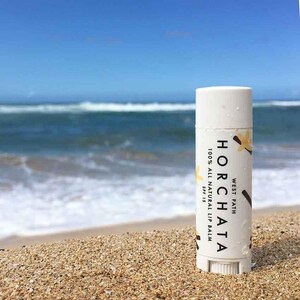 May include: A white tube of lip balm with the text "WEST PATH HORCHATA 100% ALL NATURAL LIP BALM SPF 15" on the label. The tube is sitting on a sandy beach with a blue ocean in the background.