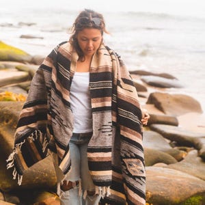 May include: A person wearing a brown, black and white striped blanket with fringe. The blanket has a geometric pattern. The person is standing on a rocky beach.