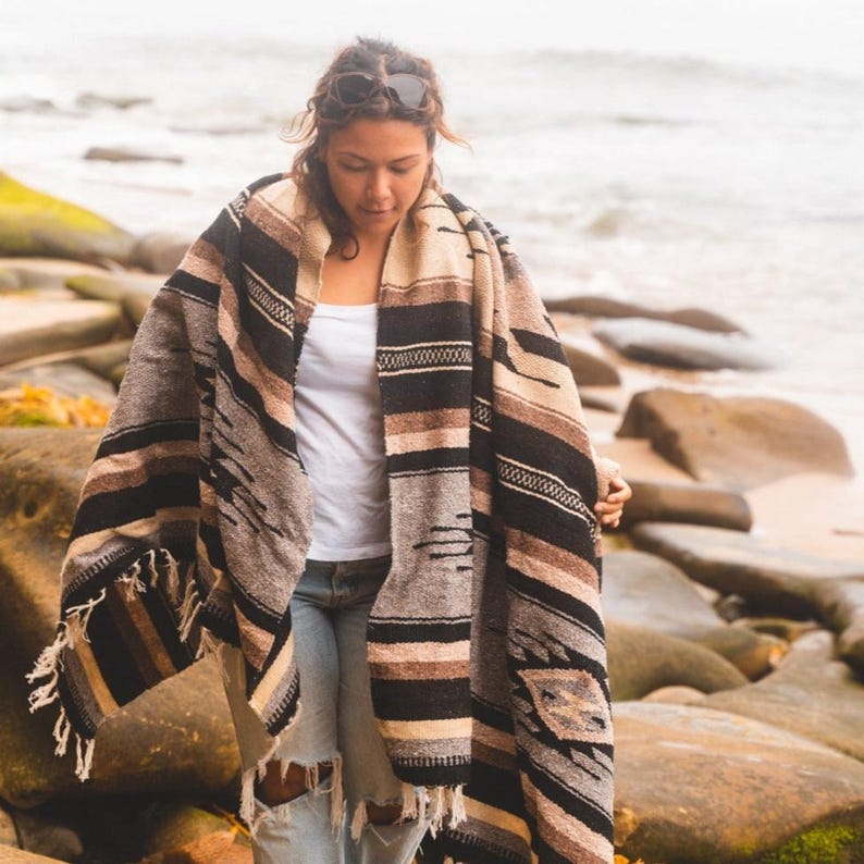May include: A person wearing a brown, black and white striped blanket with fringe. The blanket has a geometric pattern. The person is standing on a rocky beach.