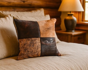 May include: Square decorative pillow with a patchwork design. The pillow features four sections of cowhide in shades of brown and tan. The pillow is placed on a beige bedspread, with a wooden headboard and a lamp in the background.