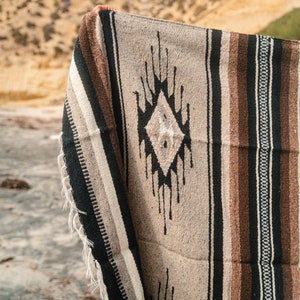 May include: A brown, black and white striped woven blanket with a black diamond pattern. The blanket has fringe on the edges.
