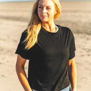 K&ouml;nnte beinhalten: Ein schwarzes T-Shirt mit Rundhalsausschnitt und kurzen &Auml;rmeln. Das T-Shirt hat eine lockere Passform. Das Model tr&auml;gt das Shirt an einem Strand. Das Shirt ist aus einem weichen, bequemen Material.