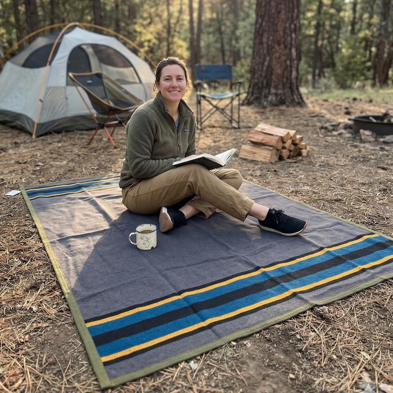 May include: A gray and blue striped outdoor blanket with a green border is spread on the ground. A person is sitting on the blanket, reading a book, with a mug of coffee nearby. A tent and camping chair are in the background.